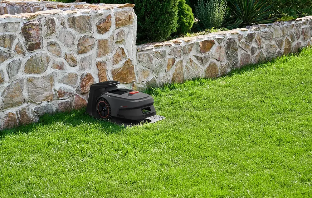 Zāles pļāvējs robots lādējas uzlādes stacijā, kas novietota blakus akmens sienai uz zāles